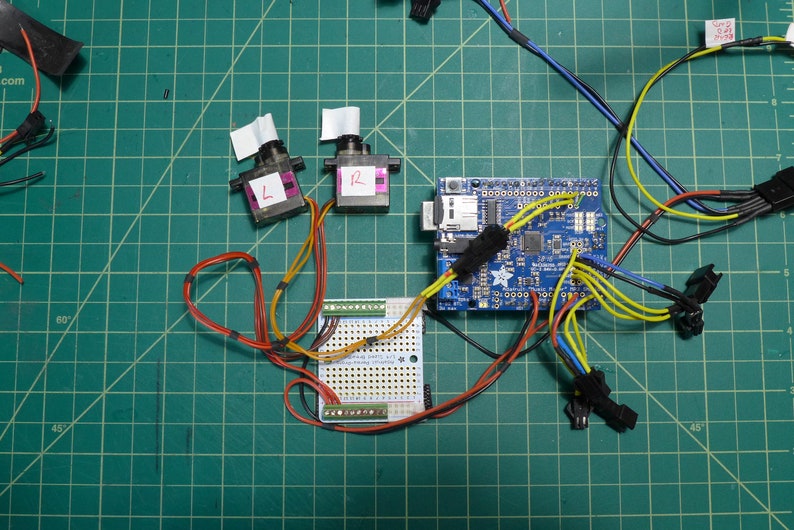 May include: A blue circuit board with wires connected to two small black motors labeled "L" and "R". The motors are connected to a breadboard with a yellow and orange wire.