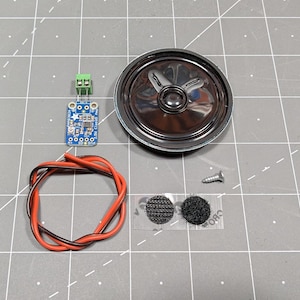 May include: An assortment of electronic components, including a small speaker, a circuit board, a screw, a pair of hook-and-loop fasteners, and a bundle of red and black wires. The components are arranged on a gray grid surface.