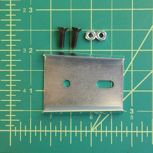 May include: A set of hardware components, including a silver metal bracket with two holes, two black screws, and two silver nuts. The items are arranged on a green grid surface.