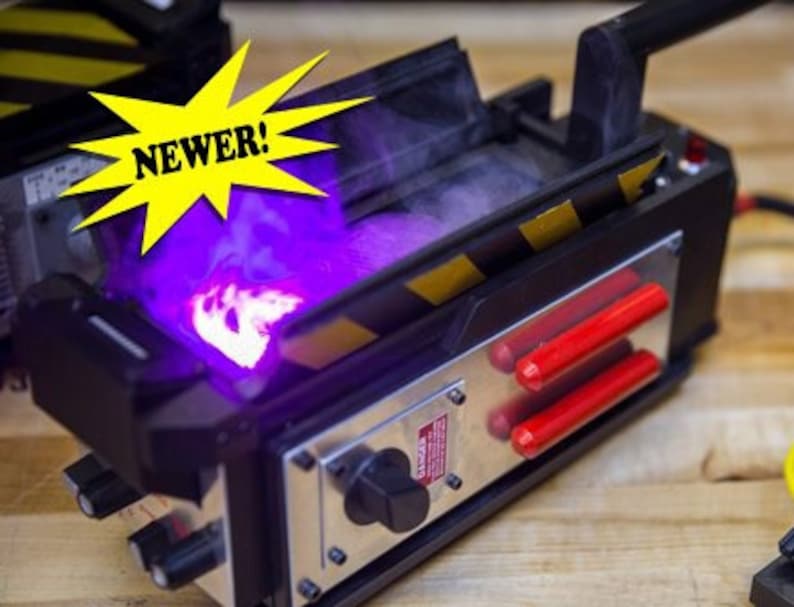 May include: A black and yellow Ghostbusters proton pack with a purple light emanating from the top. The pack has a red handle and a black and yellow striped design. The text "NEWER!" is visible in a yellow starburst.