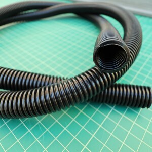 May include: A flexible black corrugated conduit tubing with a hollow cylindrical interior. The plastic hose features a continuous ribbed exterior surface designed for protective cable management and routing, coiled in a circular loop shape.