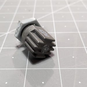 May include: A gray metal electrical component with a hexagonal shape and a series of ridges on the side. The component has a small hole in the center.