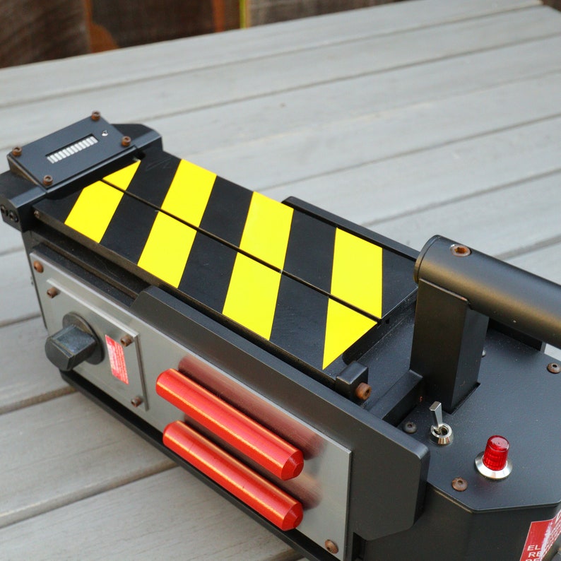 May include: A black and yellow Ghostbusters proton pack replica with a red button and a silver control panel. The pack has a black handle and a yellow and black striped design.