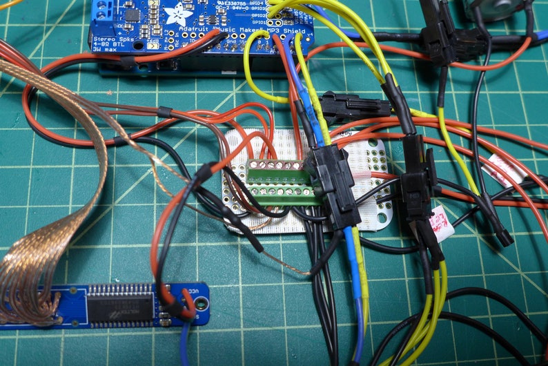 May include: A close-up of a circuit board with various wires and connectors. The wires are color-coded, including red, black, blue, yellow, and green. The circuit board has a green terminal strip with multiple pins. The wires are connected to the terminal strip and other components on the board.