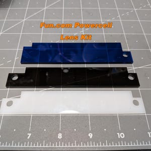 May include: Three rectangular lens pieces in blue, black, and white are arranged on a gray cutting mat. The blue lens has a patterned design. The text "Fun.com Powercell Lens Kit" is visible.