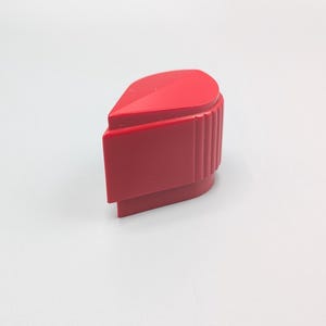 May include: A bright red plastic control knob featuring a teardrop-shaped top profile, a smooth flat front face, and a vertically ribbed side grip section. The component has a solid, molded construction with a distinct angular ridge along the top.