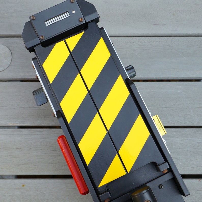 May include: A black and yellow striped toy proton pack with a red button and a small silver dial. The pack is made of plastic and has a textured surface.