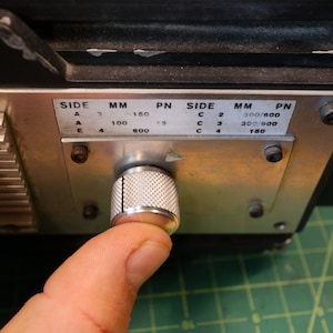May include: Close-up of a silver knob with a textured surface. The knob is labeled with the text "SIDE MM PN" and has a series of numbers and measurements. The knob is set to "600".
