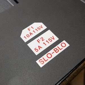 May include: A matte black surface featuring three white rectangular labels with red text. The labels read F1 10A 115V, F2 5A 115V, and SLO-BLO. The top label has a clipped corner design, while the other two are standard rectangles.