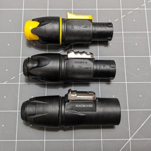 Könnte beinhalten: Drei schwarze elektrische Steckverbinder mit gelben und silbernen Akzenten. Die Steckverbinder tragen den Text "JET" und die Modellnummern "NAC3FX-W", "RCAC3M-X-000" und "JET", die darauf gedruckt sind.