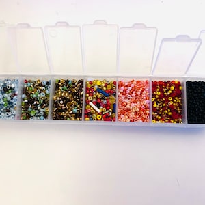 May include: A clear plastic storage box with eight compartments filled with various colours of seed beads. The beads are in the following colours: light blue, green, gold, red, orange, black, and white.