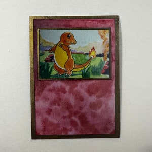 May include: Watercolor painting of a Charmander Pokémon card. The card features an orange Charmander with a yellow belly, standing in a grassy field with a flame on its tail. The card has a gold border and a red watercolor background.