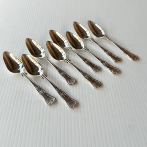 May include: Eight silver spoons displayed on a white surface. Each spoon has a rounded bowl and a decorative handle with an ornate floral design. The spoons are arranged diagonally. The reflective spoons show the surrounding environment.