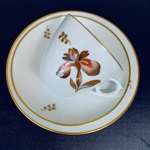 May include: White teacup and saucer set with gold trim. The teacup features a brown, orange, and purple floral design. The saucer and cup have gold accents and a simple gold leaf design.