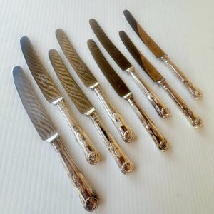 以下が含まれることがあります： 斜めに配置された6本の銀色のナイフのセット。各ナイフは、波状のテクスチャーを持つ金属製の刃と、ベースにエンボス加工された貝殻のモチーフ、ステムに沿った複雑なスクロール模様が施された装飾的なハンドルが特徴です。