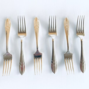 May include: Six silver forks with decorative handles, arranged on a white surface. The handles have a pointed end with a chevron pattern. The tines are evenly spaced. The forks are made of silver.