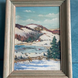 May include: A framed landscape painting shows a winter scene with snow-covered hills, a red-roofed house, and a frozen lake. The sky is light blue with white clouds. The frame is made of light, weathered wood.