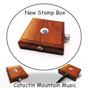 May include: A wooden stomp box with a silver knob and a jack input. The box has a googly eye on top and the text "New Stomp Box" is in a circle above the box. The text "Catoctin Mountain Music" is below the box.