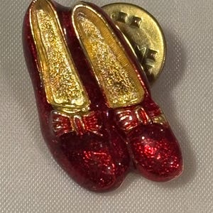 May include: A lapel pin shaped like a pair of glittery red shoes with gold-toned interiors and small gold bows on the front. The pin features a metallic gold backing with a circular clutch mechanism visible behind the shoes.