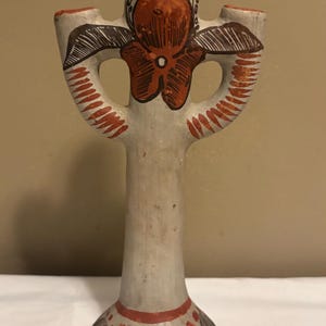 May include: A hand-painted ceramic cross-shaped candle holder. The off-white base is adorned with red and brown floral designs. The cross arms feature a floral design in red and brown. The top and base have red accents.