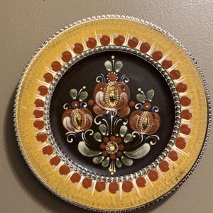 May include: Decorative ceramic plate with a yellow rim and a dark center. The plate features a floral design in shades of brown, red, green, and gold. The rim is decorated with red dots and a silver-colored border. A vintage-style piece.