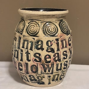 May include: A cream-colored ceramic vase with a dark rim and a textured surface. The vase features repeating spiral designs and the word "imagine" in a block letter style. The text is in black and blue.