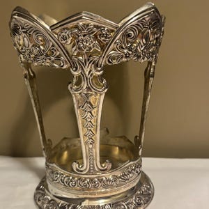 May include: A silver-toned metal glass holder featuring an intricate openwork design with embossed floral motifs and scrollwork patterns. The cylindrical structure has a scalloped top edge and a wide, decorative base with detailed engravings.