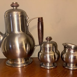 May include: A silver tea set with a teapot, creamer, and sugar bowl. The teapot has a wooden handle and a silver lid. The creamer and sugar bowl have silver lids.