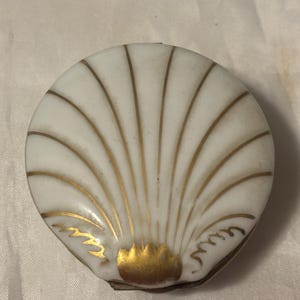 May include: A white, shell-shaped trinket box with gold accents. The box features a detailed fan design with gold lines radiating from the base. The edges are trimmed in gold, and the box is closed.