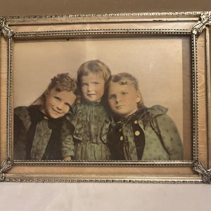 May include: Antique framed photograph featuring three children in vintage clothing. The frame is ornate with a silver finish and a light wood-tone border. The children are in a sepia tone, with hints of color in their clothing.