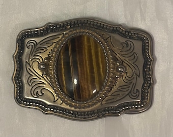 May include: Silver-toned belt buckle with an oval, tiger's eye stone center. The buckle has an ornate, engraved design around the stone and along the edges. A classic Western-style accessory.