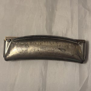 May include: A vintage silver harmonica with the text "THE DOMINATOR" and "MADE BY M. HOHNER" embossed on the cover. The harmonica has a curved shape and a metallic finish, suggesting it is made of metal.
