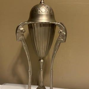 May include: A silver-colored metal incense burner with a domed lid and ornate handles. The burner has a fluted body and stands on three curved legs. The lid is topped with a decorative finial. The overall design is vintage and elegant.