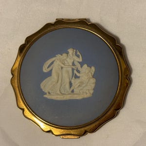 May include: A gold-toned compact with a blue enamel lid featuring a white cameo of three figures. The cameo depicts three women in classical dress, with one holding a flower.
