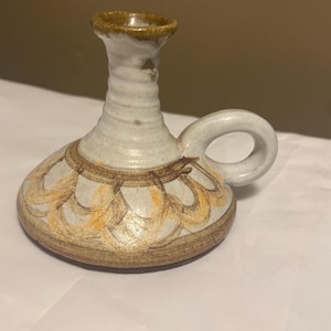 May include: A white ceramic candle holder with a brown handle and a brown and orange design. The holder has a wide base and a narrow neck.