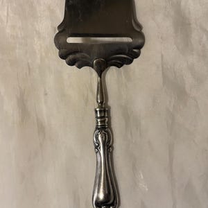 May include: A silver-toned metal cheese slicer featuring a wide, rounded blade with a horizontal cutting slot and an ornate, decorative handle with intricate embossed detailing. The tool has a polished finish and a distinct transition between the handle.