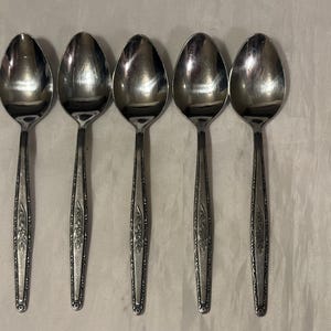 May include: Five stainless steel spoons with oval bowls and ornate, floral-patterned handles. The spoons are arranged in a row, reflecting light. The silverware is likely for dining or serving.