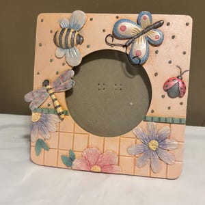 May include: Square picture frame with a peach-colored background, decorated with a bee, butterfly, dragonfly, and ladybug. The frame also features floral accents and a grid pattern, creating a whimsical, nature-inspired design.