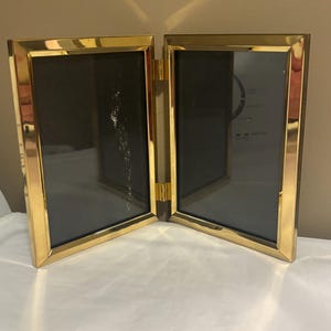 May include: A hinged, gold-toned metal double picture frame. Each frame has a black interior and a rectangular shape. The frame is open, displaying the two sides connected by a hinge. The frame is sitting on a white surface.