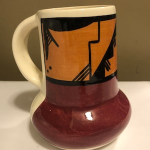 May include: A ceramic mug with a white handle and a burgundy base. The mug has a geometric design in black, orange, and white.