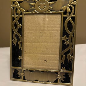 May include: A gold-toned metal picture frame with a black background. The frame features a smiling sun, stars, and clouds.