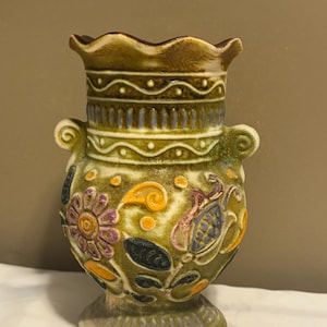 May include: A ceramic vase with a scalloped rim and two side handles. The body features a textured olive green glaze with embossed floral and leaf patterns in shades of purple, orange, and dark blue, accented by horizontal wavy line details.