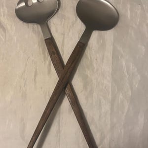 May include: A pair of salad servers with brushed stainless steel heads and long, slender dark brown wooden handles. The set includes one slotted fork and one solid spoon, arranged in a crossed position to display the metal and wood construction.