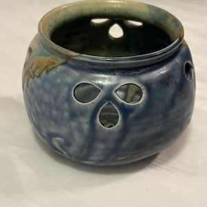 May include: A blue ceramic bowl with decorative cut-out holes. The bowl has a rounded shape with a slightly flared rim. The exterior is a mottled blue with hints of yellow and green. The bowl is likely handmade and has a rustic aesthetic.