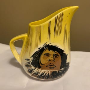 May include: Yellow ceramic creamer with a black and brown illustration of a person's face with a fur-lined hood. The creamer has a handle and a spout.