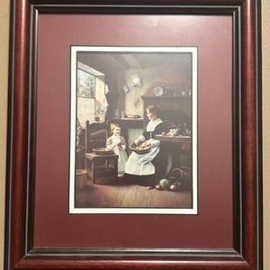 May include: Framed print depicting a woman and child in a kitchen setting. The woman, wearing a white apron, holds a bowl. The print is framed in a dark brown wood frame with a burgundy matting.