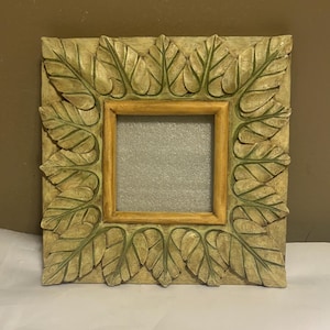 May include: Square picture frame with a leaf design. The frame is painted in a light beige color with green accents on the leaves. The inner frame is a light gold color.