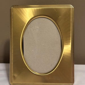 May include: A rectangular gold-colored picture frame with an oval opening. The frame has a brushed metal finish and a series of thin, parallel lines around the edges. The interior of the frame is a neutral gray color.