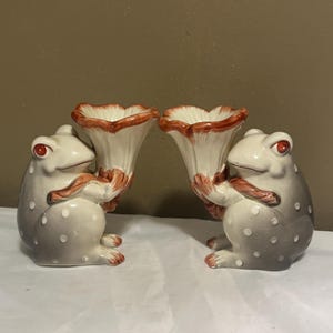 May include: A pair of ceramic frog figurines, each holding a large, flared, trumpet-shaped flower vase. The frogs are grey with white polka dots, orange eyes, and orange accents on their limbs, while the vases are cream with orange rims.