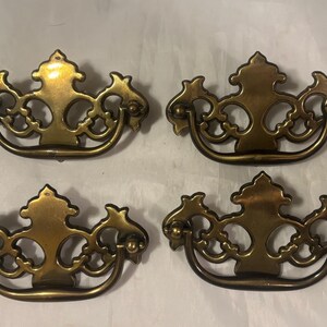May include: Four antique brass drawer pulls with ornate, symmetrical designs. Each pull features a curved handle and decorative flourishes, perfect for vintage furniture restoration. The brass has a warm, aged patina.